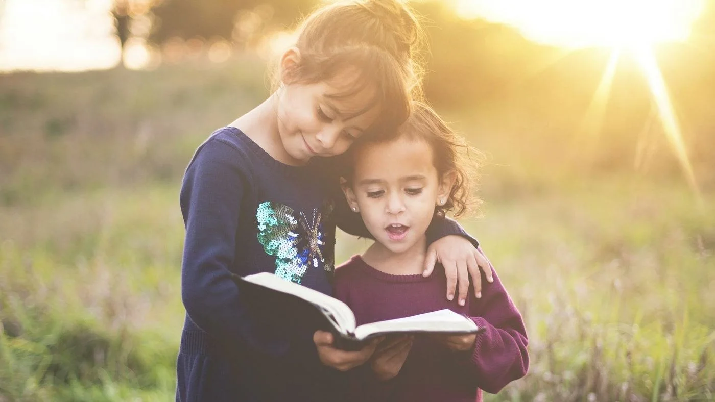 Duas irmãs leem juntas um livro aberto num campo, ao pôr do sol