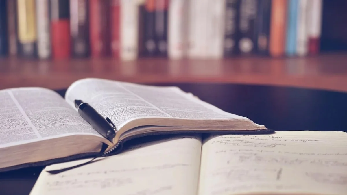 Livro aberto com uma caneta sobre as páginas e um caderno de anotações à frente, com estantes de livros ao fundo