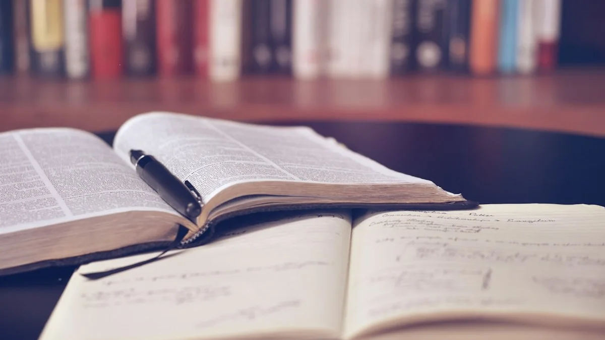 Livro aberto com uma caneta sobre um caderno de anotações escritas à mão, diante de uma estante de livros