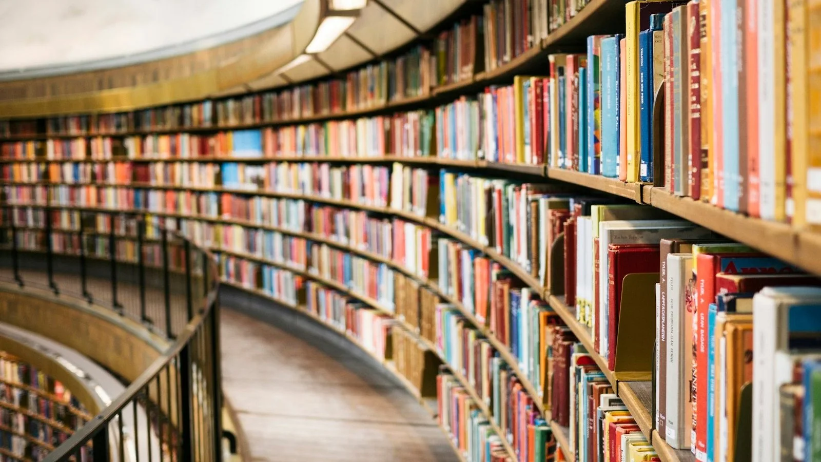 Estante curva de biblioteca, repleta de livros coloridos, com corrimão e luminárias no teto